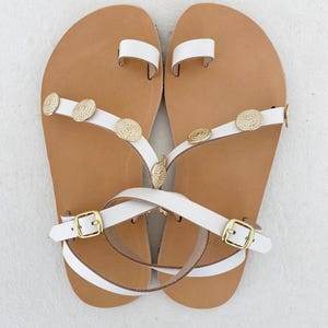 May include: A pair of white leather sandals featuring a toe loop, thin straps adorned with circular gold-toned metal embellishments, and adjustable ankle straps with gold-toned buckles. The footbeds are tan with a visible size 36 imprint.
