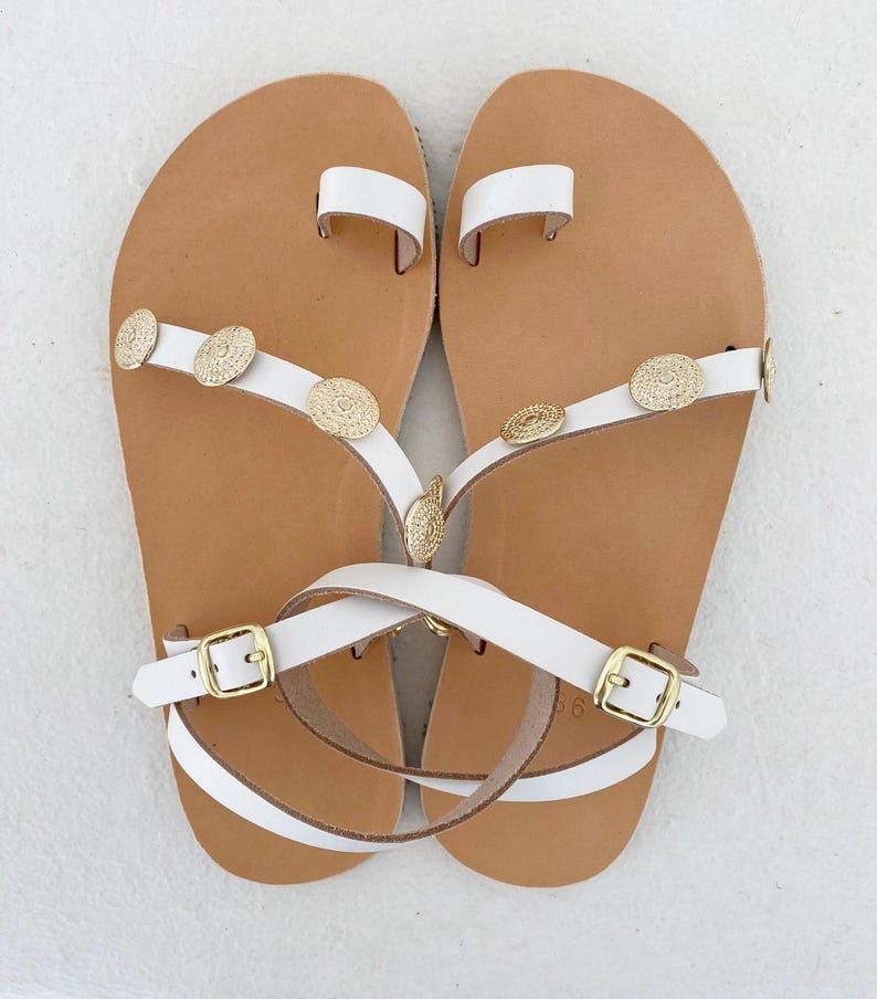May include: A pair of white leather sandals featuring a toe loop, thin straps adorned with circular gold-toned metal embellishments, and adjustable ankle straps with gold-toned buckles. The footbeds are tan with a visible size 36 imprint.