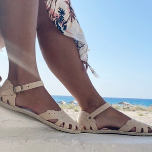 May include: A pair of cream-colored flat sandals featuring a closed-toe design with decorative cutouts, a scalloped edge along the top, and an adjustable ankle strap secured with a small bronze-toned metal buckle on the side of each sandal.
