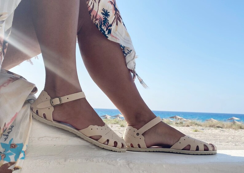 May include: A pair of cream-colored flat sandals featuring a closed-toe design with decorative cutouts, a scalloped edge along the top, and an adjustable ankle strap secured with a small bronze-toned metal buckle on the side of each sandal.