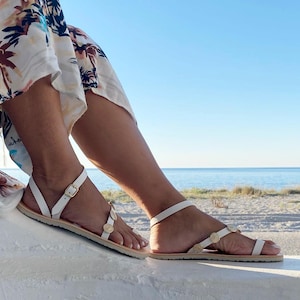 May include: A pair of white flat sandals featuring thin straps, a gold-toned buckle closure at the ankle, and small circular gold-toned decorative accents along the front strap, worn with a white skirt featuring a brown and black palm tree print.