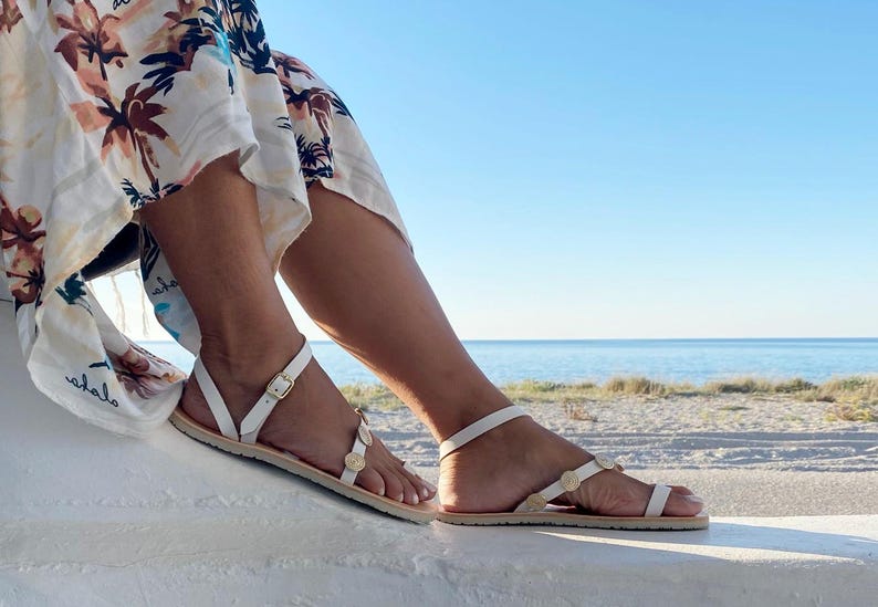 May include: A pair of white flat sandals featuring thin straps, a gold-toned buckle closure at the ankle, and small circular gold-toned decorative accents along the front strap, worn with a white skirt featuring a brown and black palm tree print.