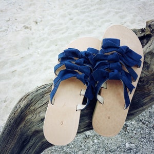 May include: A pair of blue leather sandals with multiple bow details. The sandals are on a wooden surface.