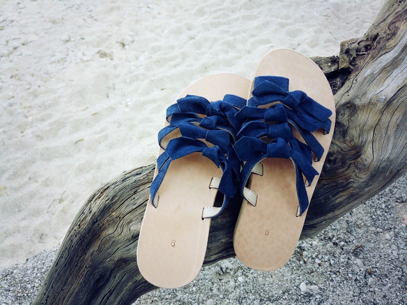 May include: A pair of blue leather sandals with multiple bow details. The sandals are on a wooden surface.