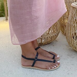 Peut inclure: Une paire de sandales en cuir noir avec une boucle et une lani&egrave;re d'orteil. Les sandales ont une semelle marron clair et sont port&eacute;es par une personne debout sur une surface de couleur claire.