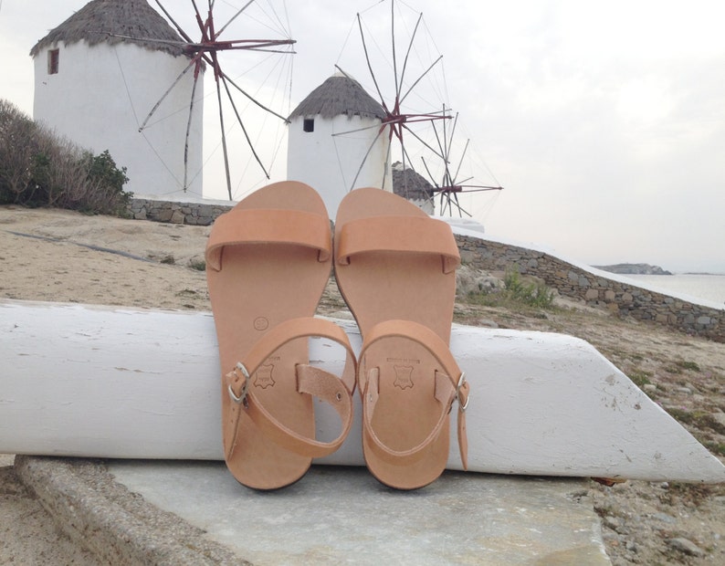 May include: A pair of tan leather sandals with a buckle closure. The sandals are sitting on a white wooden surface in front of a scenic background of windmills and a beach.