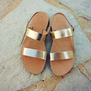 May include: A pair of flat sandals featuring two metallic gold straps across the top and a thin adjustable ankle strap with a gold buckle. The footbed is tan leather with visible stitching along the edges and the number 9 printed on the heel area.