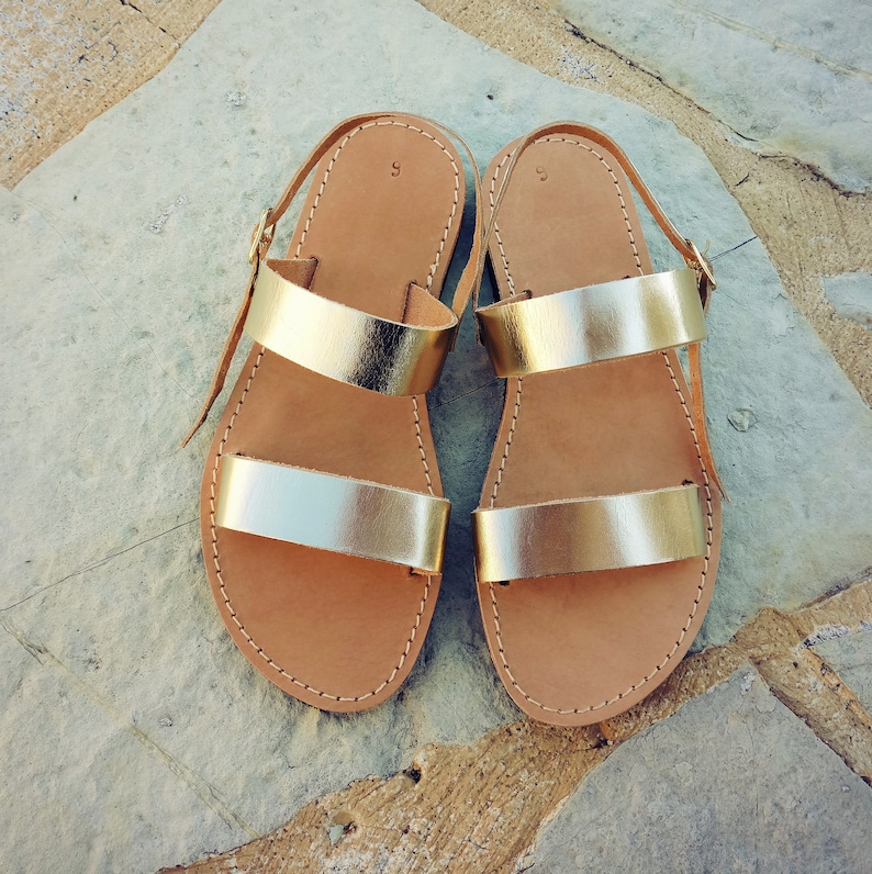 May include: A pair of flat sandals featuring two metallic gold straps across the top and a thin adjustable ankle strap with a gold buckle. The footbed is tan leather with visible stitching along the edges and the number 9 printed on the heel area.