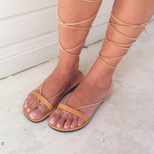 May include: Tan leather sandals with laces that wrap up the legs. The sandals have a simple design with a flat sole.