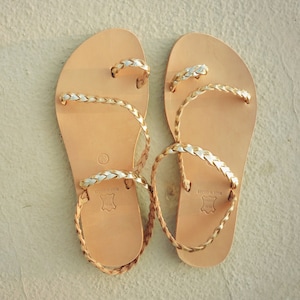 K&ouml;nnte beinhalten: Ein Paar braune Ledersandalen mit geflochtenen Riemen in Gold und Silber. Die Sandalen haben ein schlichtes Design und sind perfekt f&uuml;r den Sommer.