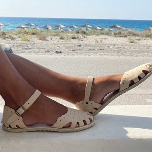 May include: A pair of beige flat sandals featuring a closed-toe design with decorative scalloped edges and cutouts. The sandals include an adjustable ankle strap with a metallic buckle and a textured sole, providing a secure fit for the wearer.