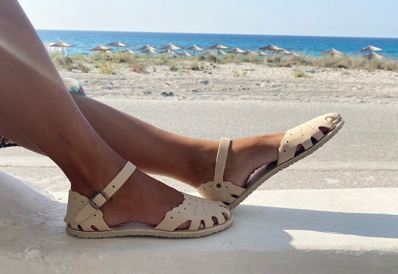 May include: A pair of beige flat sandals featuring a closed-toe design with decorative scalloped edges and cutouts. The sandals include an adjustable ankle strap with a metallic buckle and a textured sole, providing a secure fit for the wearer.