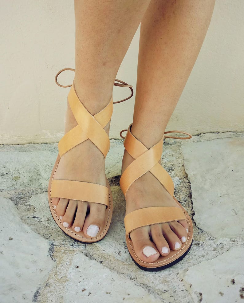 May include: A pair of light tan leather sandals featuring a horizontal toe strap and two crisscrossing straps that wrap around the ankle, secured with thin leather ties at the back. The sandals have a flat sole and a natural, un-dyed leather finish.
