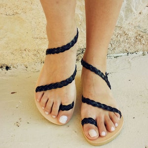 K&ouml;nnte beinhalten: Schwarze Ledersandalen mit geflochtenen Riemen, die sich um die Kn&ouml;chel und Zehen wickeln. Die Sandalen haben ein schlichtes, flaches Design und sind aus einem nat&uuml;rlichen Material gefertigt.