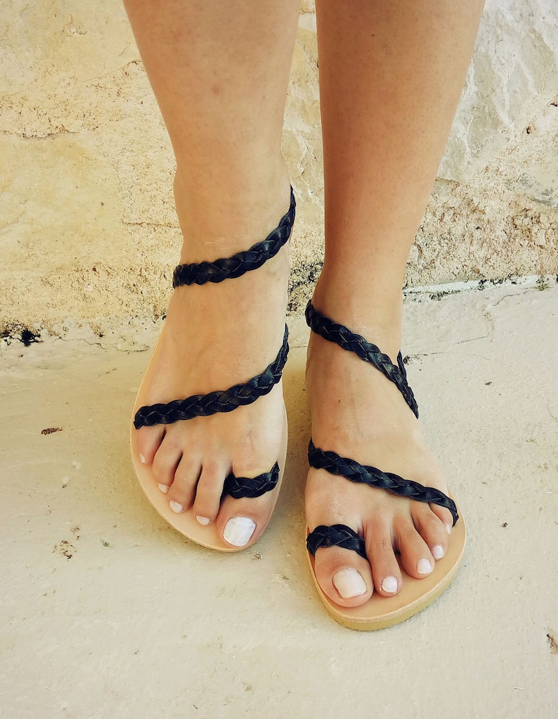 K&ouml;nnte beinhalten: Schwarze Ledersandalen mit geflochtenen Riemen, die sich um die Kn&ouml;chel und Zehen wickeln. Die Sandalen haben ein schlichtes, flaches Design und sind aus einem nat&uuml;rlichen Material gefertigt.