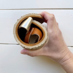 Può includere: Un contenitore in pelle arrotolato, di forma cilindrica, con un accento metallico color oro e un cinturino in pelle marrone all'interno. Il contenitore è realizzato in pelle marrone chiaro con un bordo strutturato simile alla corteccia.