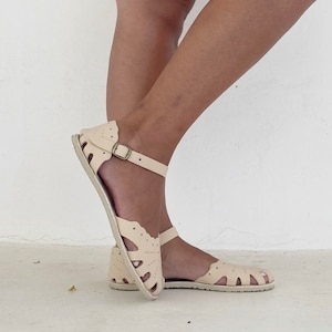 May include: Cream-colored leather sandals featuring a scalloped edge design with perforated detailing across the upper. The footwear includes an adjustable ankle strap with a metallic buckle and a flat, textured sole for a secure fit and support.