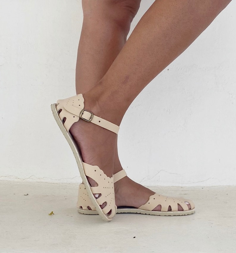 May include: Cream-colored leather sandals featuring a scalloped edge design with perforated detailing across the upper. The footwear includes an adjustable ankle strap with a metallic buckle and a flat, textured sole for a secure fit and support.