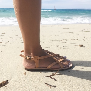 Tan leather sandals with a criss-cross strap design. The sandals are worn on a sandy beach.