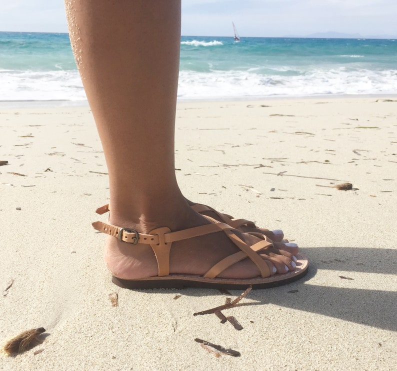 Tan leather sandals with a criss-cross strap design. The sandals are worn on a sandy beach.