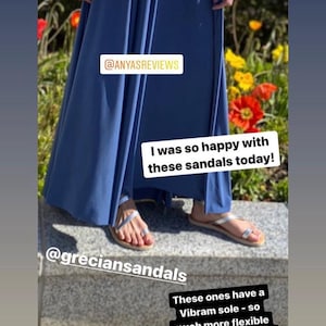 May include: Silver metallic sandals with thin straps and a toe loop, featuring a flexible Vibram sole. The sandals are worn with a long blue skirt. Text overlays include @ANYASREVIEWS, @greciansandals, I was so happy with these sandals today!, and These ones have a Vibram sole - so much more flexible than the regular ones.