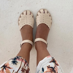 May include: A pair of cream-colored leather sandals featuring a scalloped edge, intricate perforated dot patterns across the front, and a thin ankle strap with a metal buckle closure. The sandals have a rounded toe design and a flat sole construction.