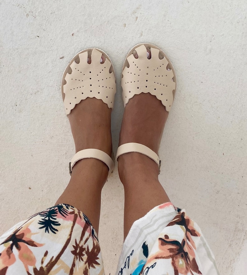 May include: A pair of cream-colored leather sandals featuring a scalloped edge, intricate perforated dot patterns across the front, and a thin ankle strap with a metal buckle closure. The sandals have a rounded toe design and a flat sole construction.
