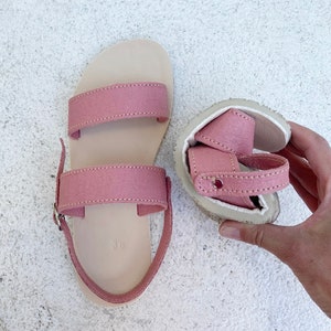 May include: A pair of dusty pink sandals featuring two wide horizontal straps across the top, a heel strap with a silver-toned metal buckle, and a light beige footbed. One sandal is laid flat, while the other is folded to show the flexible sole.