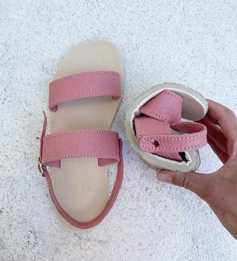 May include: A pair of dusty pink sandals featuring two wide horizontal straps across the top, a heel strap with a silver-toned metal buckle, and a light beige footbed. One sandal is laid flat, while the other is folded to show the flexible sole.