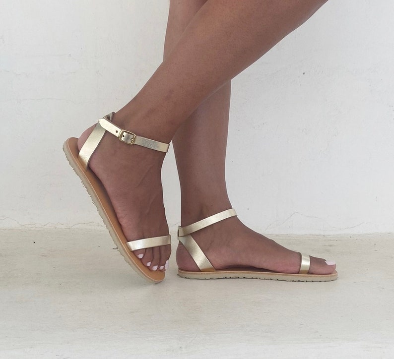 May include: A pair of gold sandals featuring thin metallic straps across the toes and an adjustable ankle strap with a small gold buckle. The sandals have a flat, light-colored sole with a textured tread, providing a simple and functional design.