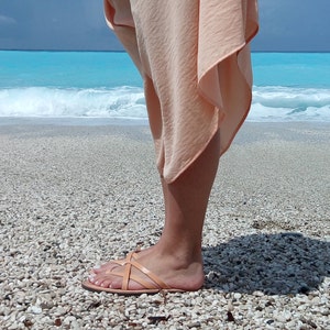 A person wearing a light peach colored skirt and sandals on a beach with white sand and blue water.