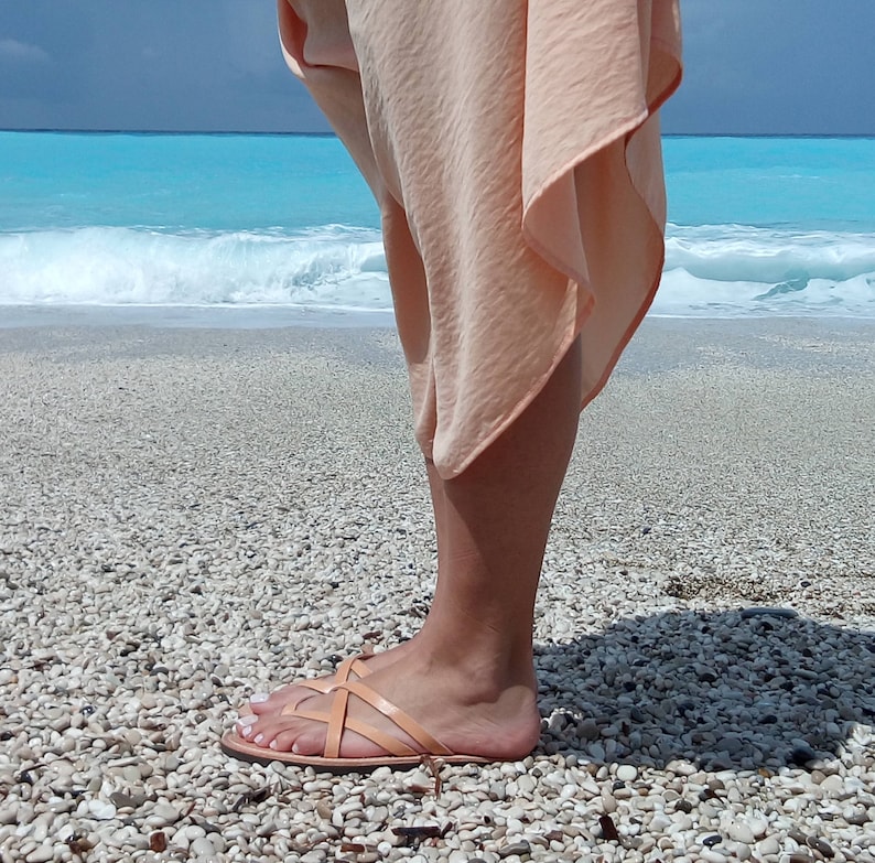 A person wearing a light peach colored skirt and sandals on a beach with white sand and blue water.