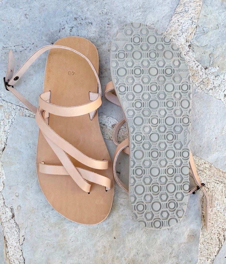 Op de afbeelding: Een paar lichtbruine leren sandalen met dunne bandjes, een verstelbare enkelgesp en een plat voetbed met de maat 40 erin gestempeld. De onderkant van de zool heeft een grijs rubberen profiel met een herhalend patroon van zeshoeken en cirkels.