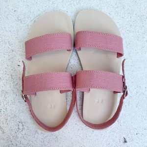 May include: A pair of flat sandals featuring two dusty rose pink straps across the top and an adjustable ankle strap with a silver metal buckle. The footbed is a light beige color and is embossed with the number 36 near the heel area.