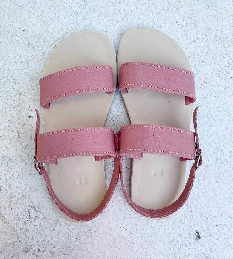 May include: A pair of flat sandals featuring two dusty rose pink straps across the top and an adjustable ankle strap with a silver metal buckle. The footbed is a light beige color and is embossed with the number 36 near the heel area.