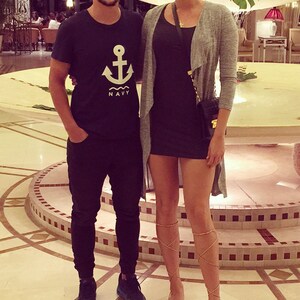 May include: A man and a woman standing in a hotel lobby. The man is wearing a black t-shirt with a white anchor and the word "NAVY" printed on it. The woman is wearing a gray cardigan over a black dress. She is also wearing brown sandals.