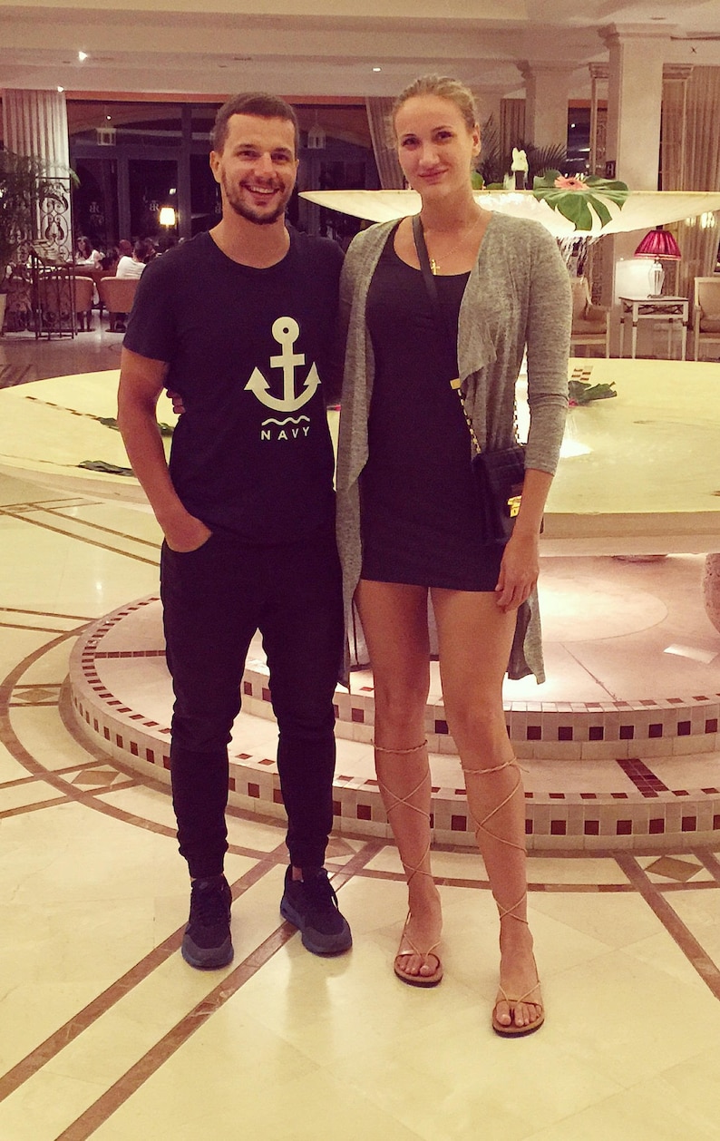 May include: A man and a woman standing in a hotel lobby. The man is wearing a black t-shirt with a white anchor and the word "NAVY" printed on it. The woman is wearing a gray cardigan over a black dress. She is also wearing brown sandals.