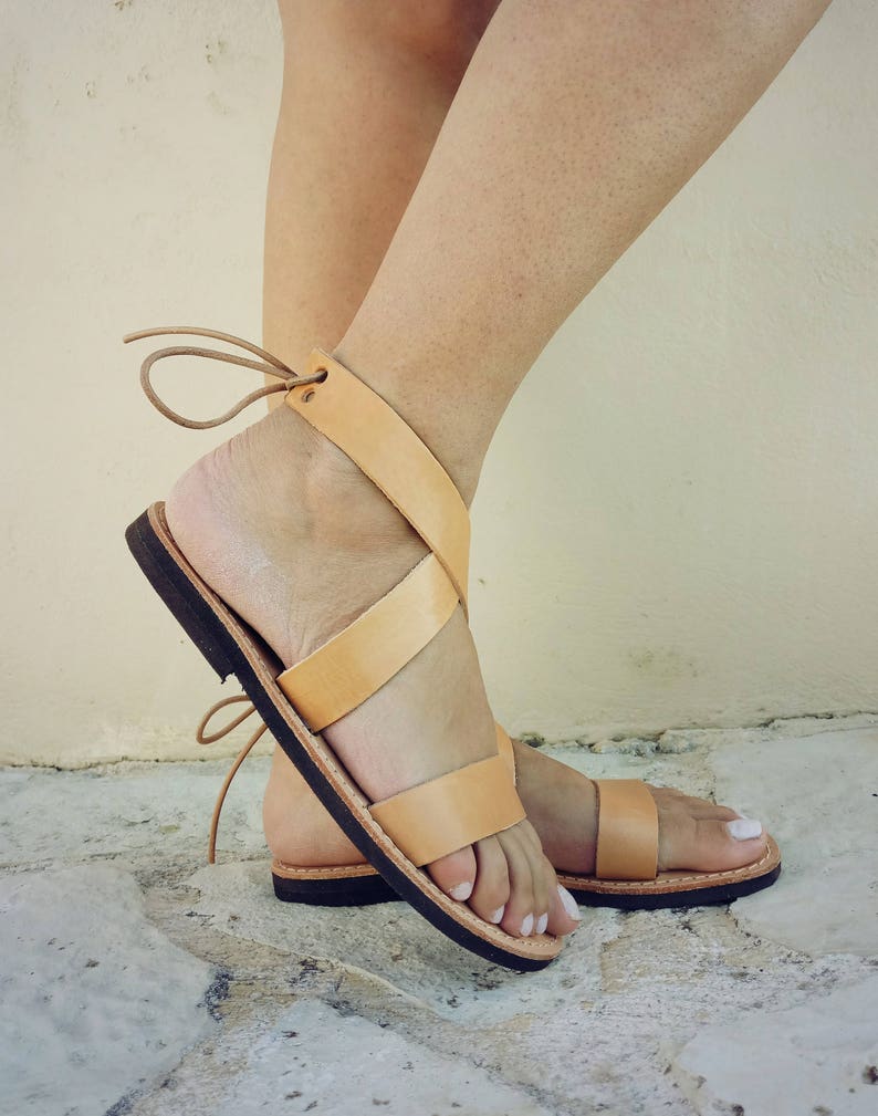 May include: A pair of tan leather sandals featuring wide straps that wrap around the ankle and secure with thin leather ties. The sandals have a flat, dark brown sole and an open-toe design, providing a simple and functional footwear option.