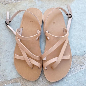 Op de afbeelding: Een paar lichtbruine leren sandalen met meerdere dunne bandjes over de voet, een teenlus en een verstelbare enkelband met een kleine metalen gesp. Het voetbed van elke sandaal is voorzien van het ingedrukte cijfer 40 voor de maat.
