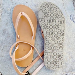 Könnte beinhalten: Ein Paar hellbraune Ledersandalen mit dünnen Knöchelriemen und Zehensteg. Eine Sandale zeigt das glatte Lederfußbett mit der eingestanzten Zahl 36, während die andere die graue Vibram-Gummilaufsohle mit einem sechseckigen Muster aufweist.