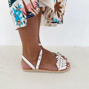 May include: White flat sandals featuring a decorative leaf-shaped strap design across the top of the foot, a thin ankle strap with a silver-toned metal buckle closure, and a light tan sole, worn with a patterned skirt featuring palm tree motifs.
