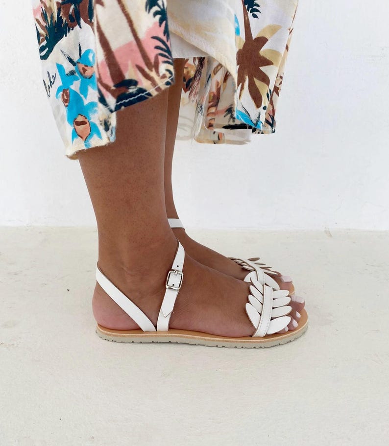 May include: White flat sandals featuring a decorative leaf-shaped strap design across the top of the foot, a thin ankle strap with a silver-toned metal buckle closure, and a light tan sole, worn with a patterned skirt featuring palm tree motifs.
