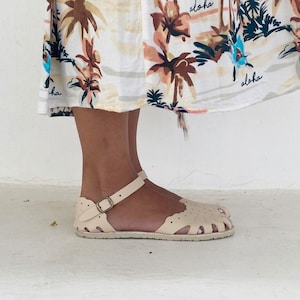 May include: A pair of cream-colored closed-toe sandals featuring a scalloped edge design, cut-out details, and an adjustable ankle strap with a metallic buckle. The sandals are worn beneath a white skirt patterned with brown palm trees and the word aloha.