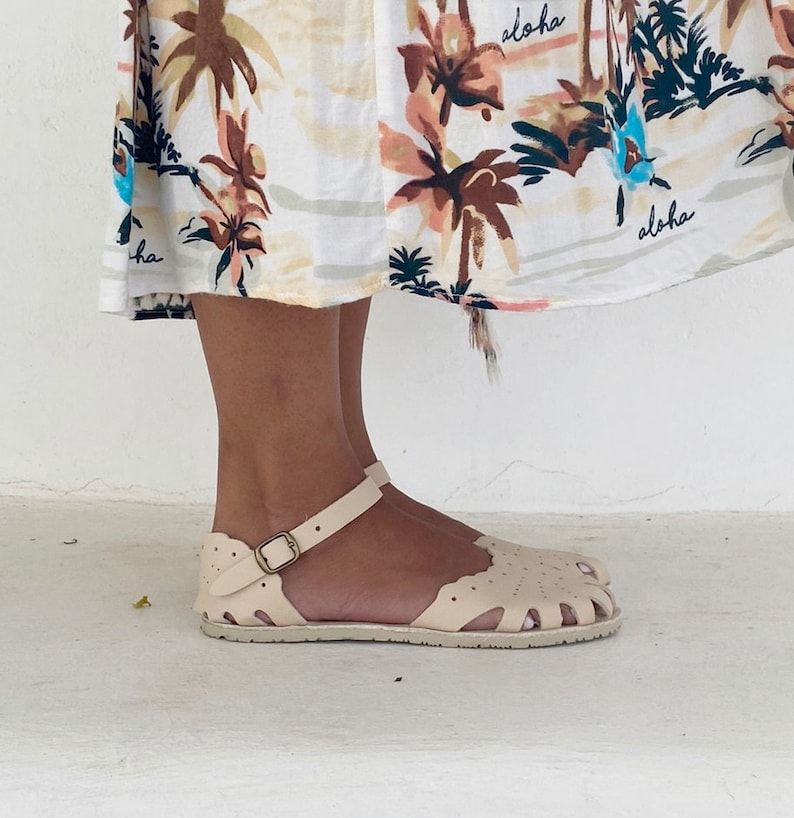May include: A pair of cream-colored closed-toe sandals featuring a scalloped edge design, cut-out details, and an adjustable ankle strap with a metallic buckle. The sandals are worn beneath a white skirt patterned with brown palm trees and the word aloha.
