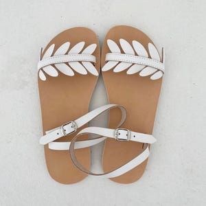 May include: A pair of flat sandals featuring a tan footbed and white leather straps. The front strap is decorated with a series of white leaf-shaped cutouts. Each sandal includes a thin white ankle strap secured with a silver-toned metal buckle.