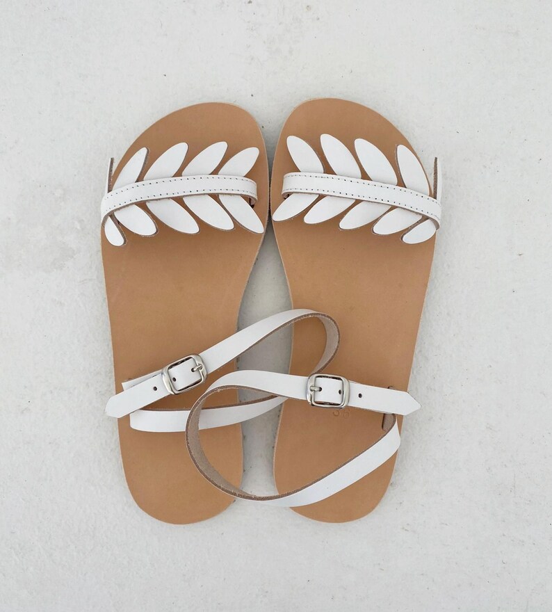 May include: A pair of flat sandals featuring a tan footbed and white leather straps. The front strap is decorated with a series of white leaf-shaped cutouts. Each sandal includes a thin white ankle strap secured with a silver-toned metal buckle.