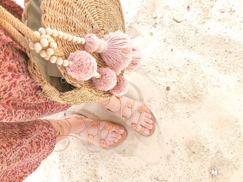 Op de afbeelding: Een ronde tas van gevlochten stro met lichtroze wollen pompons en een grote kwast, gecombineerd met beige leren vetersandalen. De tas heeft een lange gevlochten schouderband en wordt gedragen bij een jurk met een rood en bruin bloemenpatroon.