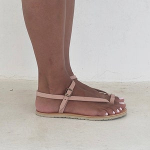 Peut inclure: Une paire de sandales en cuir rose clair avec une fermeture &agrave; boucle et une bride de style thong. Les sandales ont une semelle plate et sont faites d'un cuir souple et doux.