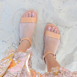 May include: A pair of light brown leather sandals with ankle straps. The sandals are on a sandy surface.