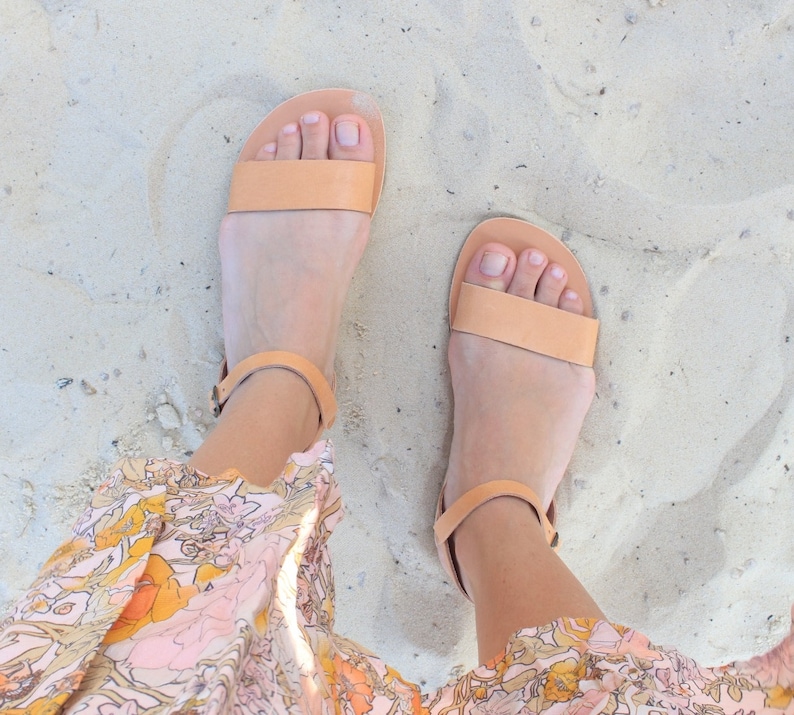 May include: A pair of light brown leather sandals with ankle straps. The sandals are on a sandy surface.